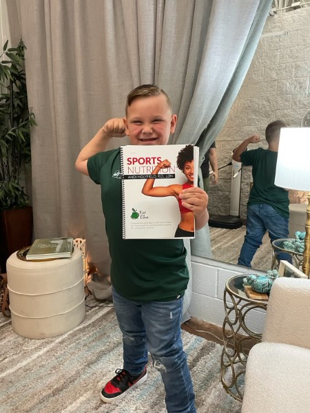 Photo of a young boy smiling and holding a copy of the book Sports Nutrition, mimicking the muscle pose on the cover after losing 26 pounds following the Eat to Lose program